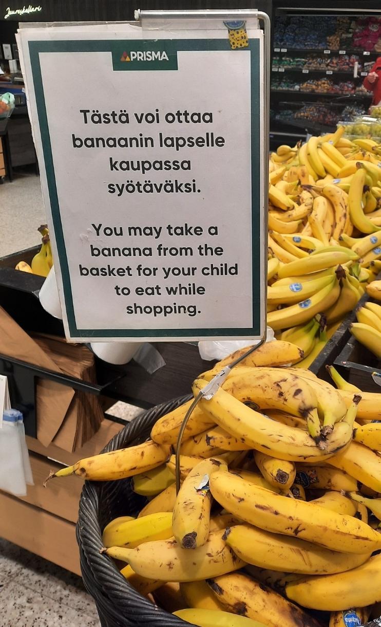 Bananen im Supermarkt. Im Vordergrund ein Korb reifer Bananen mit kleineren braunen Stellen, jedoch in einwandfreiem Zustand. Darüber ein Schild, das auf finnisch und deutsch erklärt: Die Bananen sind für Kinder zum Essen während des Einkaufs.